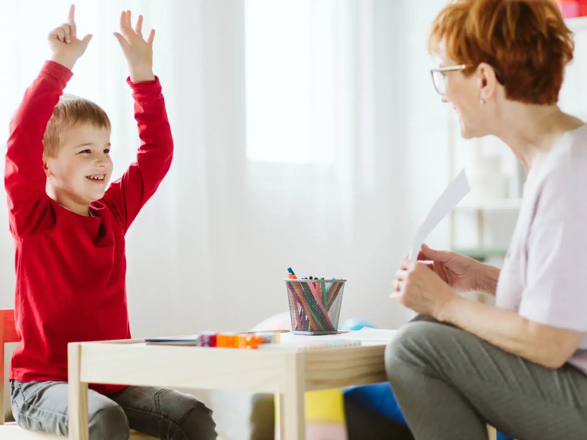 Boy-with-dyspraxia-raising-both-hands-while-speech-pathologist-holds-a-paper-during-therapy-session