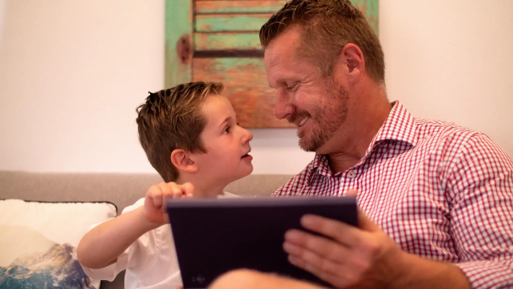 A smiling father supports his young son as they use a tablet-based speech-generating device together at home, showcasing active communication modeling used in AAC speech therapy.