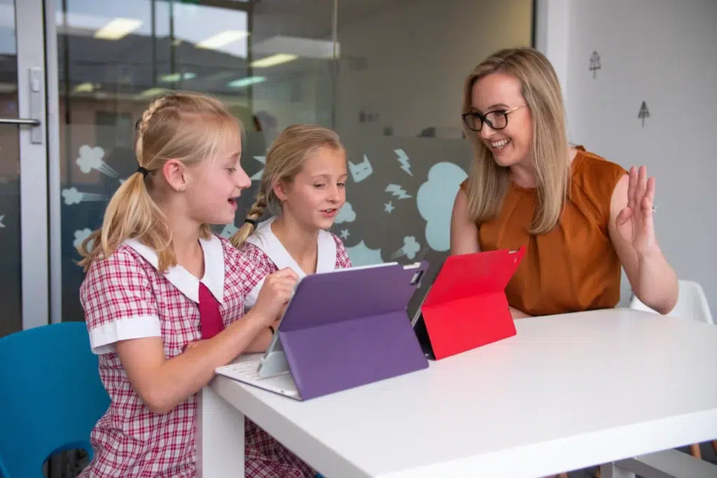 Two young girls interact with tablet communication setups during a child-led AAC speech therapy session with a female therapist.